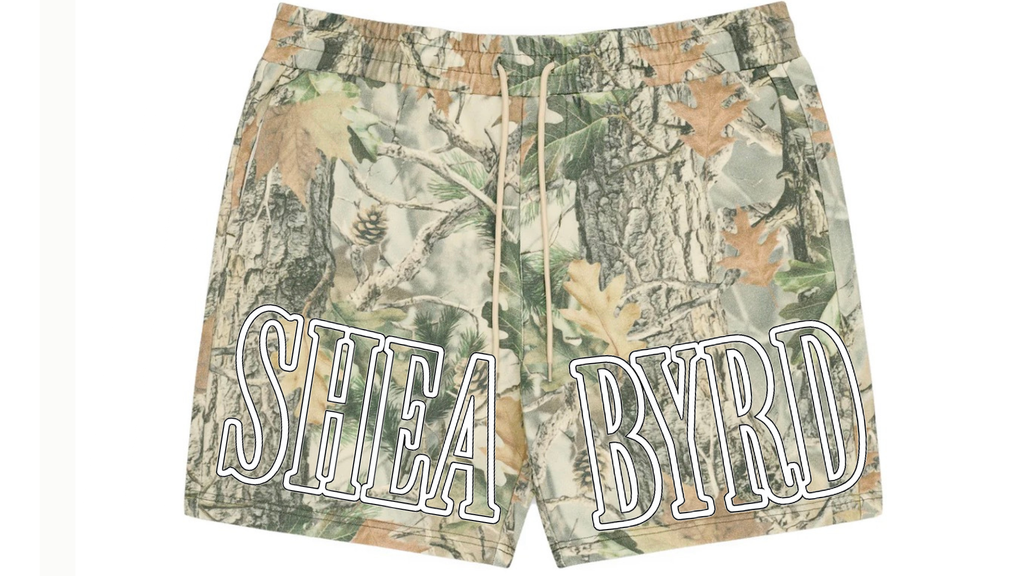A pair of camouflage print shorts featuring a woodland camo pattern in shades of green, brown, and beige, with 'SHEA BYRD' printed in large white varsity-style block letters outlined in black across the front.